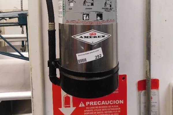 Stainless Class K fire extinguisher on a restaurant kitchen wall with a red caution sign mounted below it, photographed on a Boss Fire Protection job.